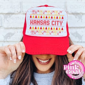 May include: A red and white trucker hat with the text "KANSAS CITY" in red. The hat features a festive design with footballs, presents, and Christmas trees. The hat has a red brim and a white mesh back.