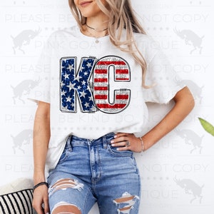 May include: White t-shirt with a glittery red, white, and blue "KC" design. The letters are outlined in silver and have a stitched look.
