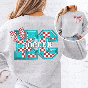 Op de afbeelding: Een lichtgrijs sweatshirt met een groot turquoise en rood geblokt ontwerp met de tekst "KC SOCCER". Een rood-witte geblokte strik staat linksboven en rechtsboven op de trui. De trui is een crew neck met lange mouwen.