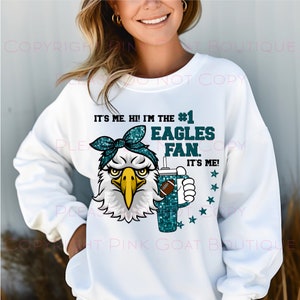 May include: White sweatshirt with a green glitter eagle wearing a blue bandana and holding a green glitter cup with a football. The text on the sweatshirt reads "It's me. Hi! I'm the #1 Eagles Fan. It's me!"