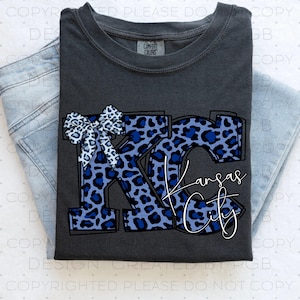 May include: Dark grey t-shirt featuring a blue leopard print "KC" design and a bow. The words "Kansas City" are written in a script font. The shirt is folded on top of a pair of light blue jeans.