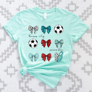 May include: A light teal t-shirt featuring various soccer-themed graphics. The design includes soccer balls, bows with different patterns, and the phrases "Kansas City" and "soccer fan". A water bottle and headphones are also included.