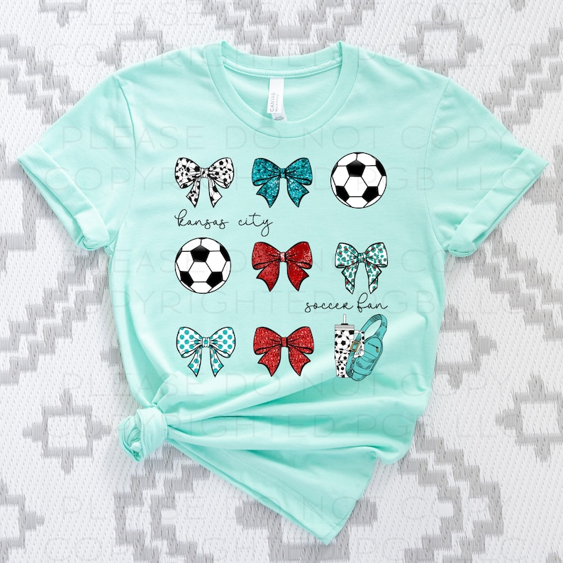 May include: A light teal t-shirt featuring various soccer-themed graphics. The design includes soccer balls, bows with different patterns, and the phrases "Kansas City" and "soccer fan". A water bottle and headphones are also included.