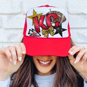 May include: Red and white trucker hat with a white mesh back. The front of the hat features a large glittery red "KC" with a gold star, a gold bow, a white star, and a football. The hat is being held up to a person's face.