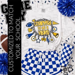 Könnte beinhalten: Ein weißes T-Shirt mit einer Grafik eines Bettelarmbands mit dem Text 'CHARMED to be a VIKING', umgeben von fußballbezogenen Anhängern, kombiniert mit blau-weiß karierten Shorts und einem passenden blau-weiß karierten Becher.
