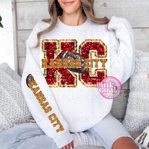 May include: White sweatshirt featuring the text "KC KANSAS CITY" in red and gold glitter. A football graphic is in the center. The sleeve also has "KANSAS CITY" in gold glitter. The sweatshirt is a fan item.