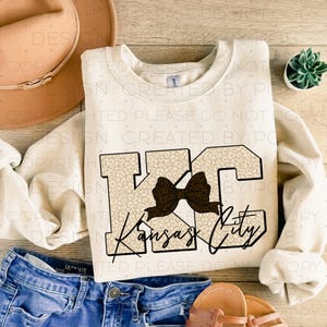 May include: A cream-coloured sweatshirt with a black and brown bow and the text "Kansas City" in black letters with a leopard print background.