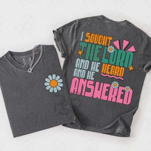 May include: Two charcoal gray t-shirts. One features a daisy graphic and a silver chain necklace. The other has the text "I SOUGHT THE LORD AND HE HEARD AND HE ANSWERED" in orange, green, and pink. The shirts are likely made of soft cotton.