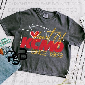 Può includere: Una maglietta grigio antracite con un design grafico. Il design presenta un contorno bianco di uno stato con un cuore rosso, le parole "Heart Of KCM" in rosso e oro, e "CREATED BY" e "SINCE 1963" in oro. La maglietta è piegata.