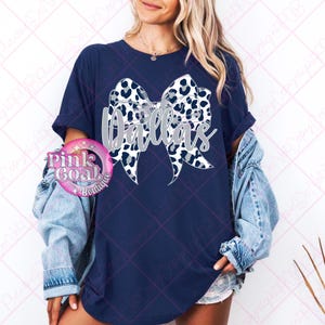 May include: A navy blue t-shirt featuring a white and grey animal print bow with the word "Dallas" in silver glitter lettering.