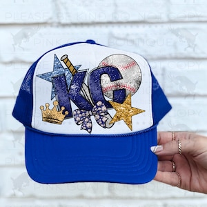 May include: A royal blue trucker hat with a white mesh back. The front of the hat features a white background with a glittery blue "KC" with a baseball, a star, a crown, and a bow.