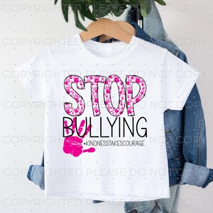 Puede incluir: Camiseta blanca con un diseño de huellas de manos rosas que dice "STOP BULLYING" y "#KINDNESSTAKESCOURAGE".