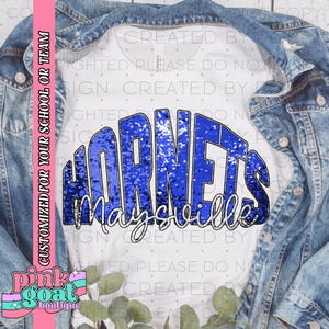 Może przedstawiać: Biała koszulka z niebieskim cekinowym wzorem "Hornets Maysville". Koszulka jest prezentowana na dżinsowej kurtce. Tekst "CUSTOMIZED FOR YOUR SCHOOL OR TEAM" znajduje się po lewej stronie obrazu.