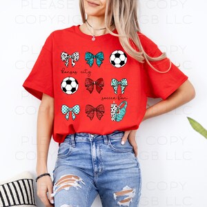 May include: A red t-shirt featuring a soccer theme. The shirt displays soccer balls, bows, and the text "Kansas City" and "soccer fan". The bows have various patterns and colors, including black and white, teal, and red.
