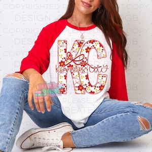 May include: A white and red raglan shirt featuring the text "KC Kansas City" in a floral design. The letters "KC" are filled with red flowers and yellow dots. The shirt has red sleeves and a white body.
