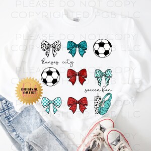 May include: White sweatshirt featuring soccer-themed graphics. The design includes soccer balls, bows in various patterns and colors, and the text "Kansas City" and "soccer fan". The sweatshirt also has a "Original Artist" seal.