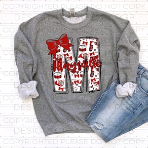 Puede incluir: Una sudadera gris con una gran letra "M" y la palabra "Maysville" en escritura roja. La "M" está llena de gráficos de instrumentos musicales y un lazo rojo. La sudadera se combina con unos vaqueros azules.