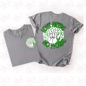 Op de afbeelding: Twee grijze t-shirts met de tekst "LOVE NEEDS NO WORDS" in het groen. Eén shirt heeft het ontwerp op de voorkant, de andere op de zak. Het ontwerp bevat een handgebaar, groene harten en een groen lint.