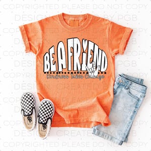 Puede incluir: Camiseta naranja con las palabras "BE A FRIEND" en negrita, fuente blanca, con un gráfico de gesto de la mano. La frase "Kindness Takes Courage" está debajo del texto principal. La camiseta se combina con pantalones cortos de mezclilla y zapatos sin cordones a cuadros.