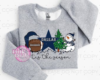 Puede incluir: Sudadera gris con un diseño festivo. Presenta un balón de fútbol con gorro de Papá Noel, una estrella azul con el texto "DALLAS", un árbol de Navidad y un muñeco de nieve. El texto "tis the season" está debajo. Un logotipo de Pink Goal Boutique.