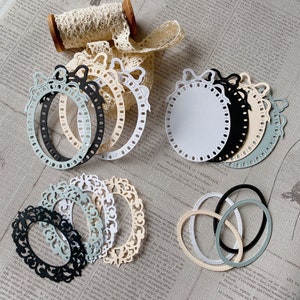 Op de afbeelding: Assortiment van ovale borduurringen in wit, zwart en beige met decoratieve strikken. De ringen zijn in een cirkelvormige opstelling op een wit en grijs gepatroneerd oppervlak.