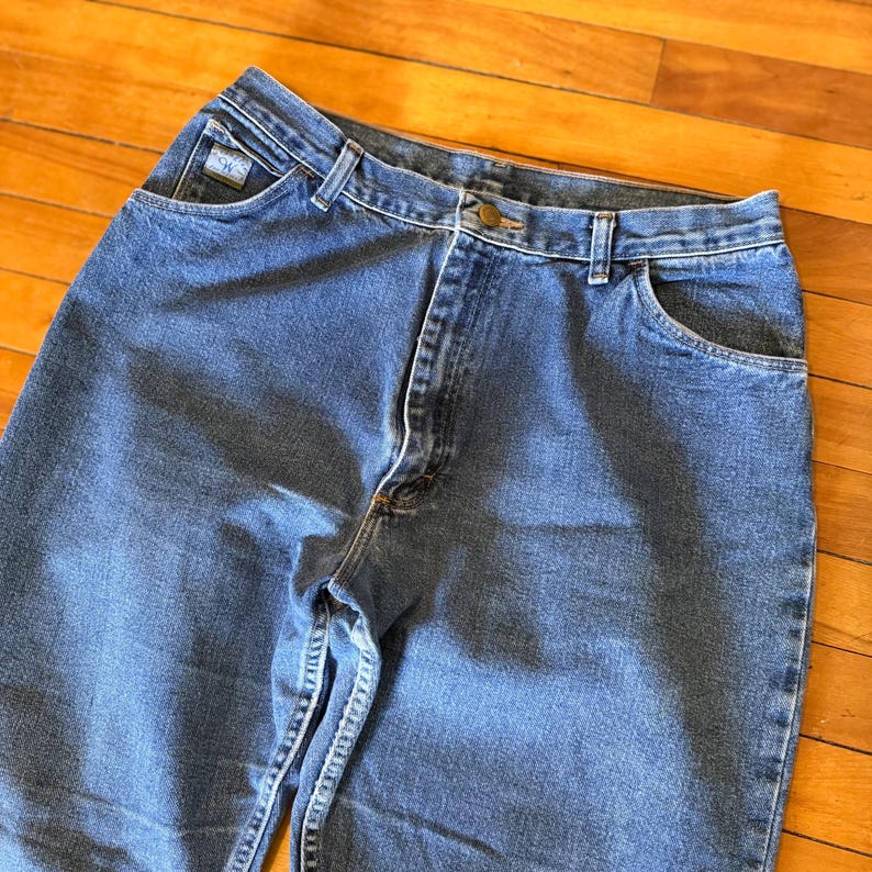 May include: Close-up of blue denim jeans. The jeans have a classic five-pocket design, a button and zipper closure, and belt loops. The jeans have a faded appearance and a small logo on the front pocket.