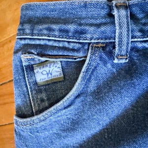 May include: Close-up of blue denim jeans with a pocket and a small label that reads "Wrangler for women". The jeans have a classic design with visible stitching and a button closure.