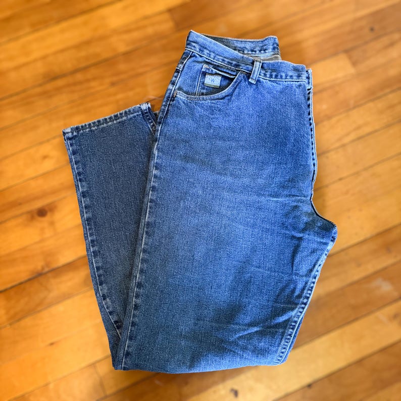 May include: A pair of blue denim jeans with a classic five-pocket design. The jeans have a straight leg cut and a button and zipper closure. The jeans are made of a medium-wash denim fabric and feature a small logo on the front pocket.
