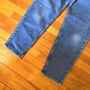 May include: Close-up of two pairs of light blue denim jeans. The jeans are laid out on a wooden surface, showcasing the details of the fabric and stitching. The jeans have a classic, casual style.
