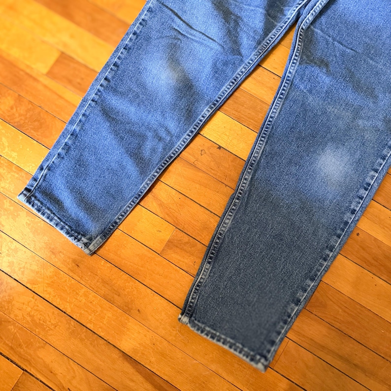 May include: Close-up of two pairs of light blue denim jeans. The jeans are laid out on a wooden surface, showcasing the details of the fabric and stitching. The jeans have a classic, casual style.