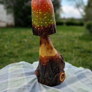 May include: A handcrafted wooden mushroom sculpture with a vibrant color scheme. The mushroom cap transitions from red to yellow and green, speckled with yellow dots. The stem and base are natural wood tones, showcasing the wood's texture.