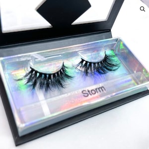 May include: A pair of false eyelashes in a clear plastic case with a black background. The case has a holographic iridescent finish and the text "Storm" is printed on the bottom.