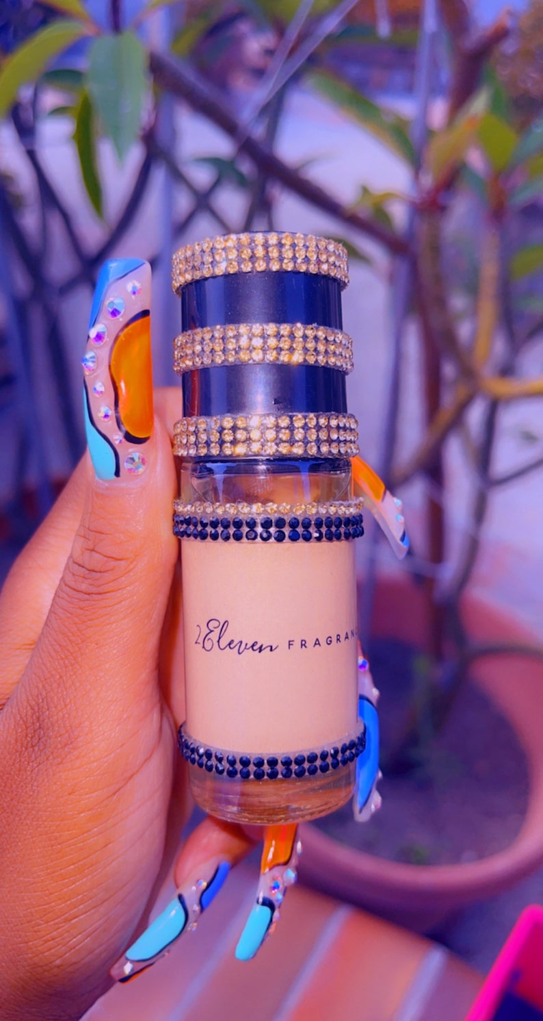 K&ouml;nnte beinhalten: Eine kleine, durchsichtige Glasflasche mit einem schwarzen und goldenen Verschluss. Die Flasche ist mit Strasssteinen verziert und tr&auml;gt den Schriftzug "2 Eleven Fragrances" auf dem Etikett.