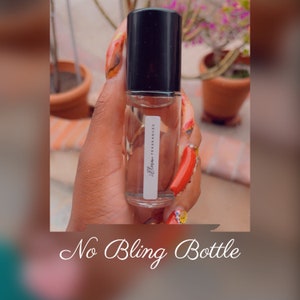 K&ouml;nnte beinhalten: Eine transparente Glas-Rollflasche mit schwarzem Verschluss und einem wei&szlig;en Etikett mit der Aufschrift "Elara Fragrances". Die Flasche wird von einer Hand mit rotem und orangefarbenem Nagellack gehalten.