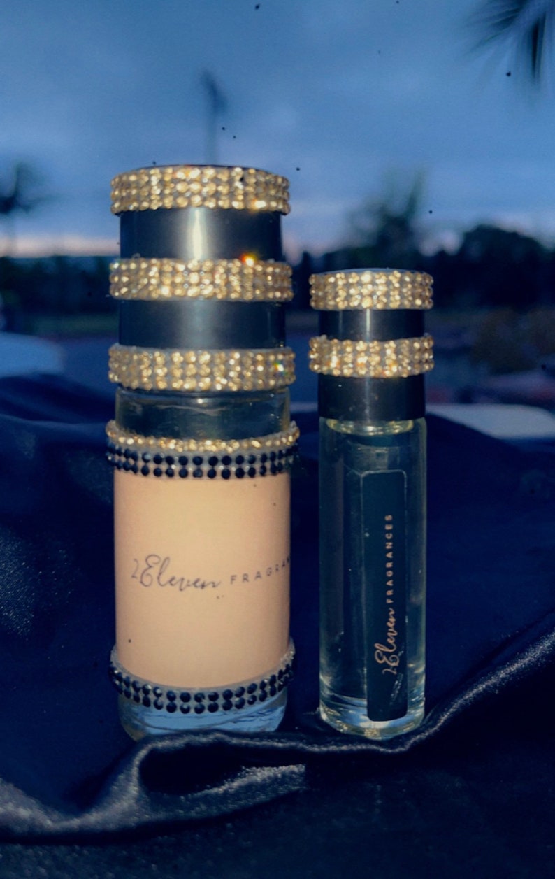 K&ouml;nnte beinhalten: Zwei Glasflaschen mit Parf&uuml;m mit schwarzen und goldenen Etiketten. Die gr&ouml;&szlig;ere Flasche hat ein schwarzes und goldenes Etikett mit dem Text "2 Eleven Fragrances". Die kleinere Flasche hat ein schwarzes Etikett mit dem Text "2 Eleven Fragrances". Beide Flaschen haben ein goldenes und schwarzes Juwelenmuster um den oberen Teil.