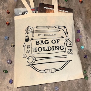 May include: Off-white canvas tote bag with black illustrations of fantasy weapons and the text "BAG OF HOLDING." The bag contains books and is surrounded by colorful dice, perfect for tabletop gaming enthusiasts.