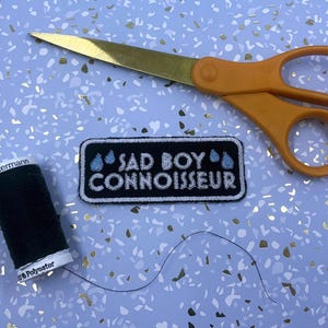 May include: An orange-handled pair of scissors with gold blades, a black patch embroidered with "SAD BOY CONNOISSEUR" and a spool of dark green thread. The items are on a light blue surface with gold flecks.