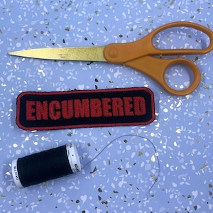 May include: An orange-handled pair of scissors with gold blades, a black and red embroidered patch with the word "ENCUMBERED", and a spool of dark green thread on a light blue background.