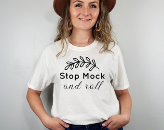 Puede incluir: Camiseta blanca con un gráfico negro que dice "Stop Mock and roll" con un pequeño diseño de hoja sobre el texto. La persona que lleva la camiseta sonríe y lleva un sombrero marrón.