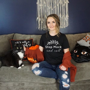Può includere: Una donna indossa una maglietta nera con la scritta "Stop Mock and roll" e un disegno a foglia. È seduta su un divano grigio con un gatto bianco e nero e cuscini a tema Halloween.