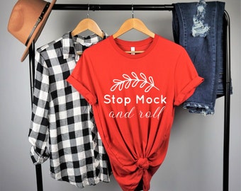 May include: Red t-shirt with white text that says "Stop Mock and roll" and a small white leaf design. The shirt is on a hanger with a black and white plaid shirt and blue jeans.