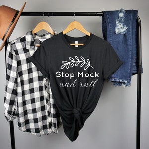 May include: A black t-shirt with white text that reads "Stop Mock and roll" and a small white leaf design. The shirt is hanging on a black rack with a plaid shirt and blue jeans.