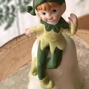 May include: A porcelain bell figurine featuring a small elf with orange hair, a green hat, and a yellow and green tunic. The elf is perched on top of the bell, which is white with a subtle green gradient and embossed floral patterns on the base.