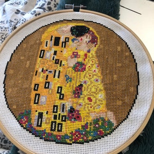 May include: A cross-stitch embroidery of Gustav Klimt's The Kiss, featuring two figures in a golden embrace, surrounded by colorful flowers. The embroidery is framed in a wooden hoop.