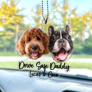 May include: A clear acrylic car charm with a chain for hanging. The charm features two dog faces, a brown and white doodle and a black and white French bulldog. The text "Drive Safe Daddy Lucas & Coco" is printed on the charm.