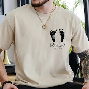 May include: A beige t-shirt with a black imprint of two baby footprints and the text "Olivia Jane APR 11, 2024".