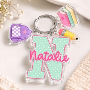 May include: A clear acrylic keychain featuring a large mint green letter N with white polka dots and the name Natalie in pink script. Attached are three smaller charms: a purple checkered backpack, a stack of three books, and a yellow pencil.