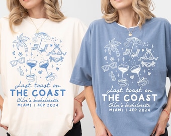 May include: Two t-shirts with a white graphic design of a beach scene with palm trees, sunglasses, an umbrella, and champagne flutes. The text on the shirts reads "Last toast on THE COAST Chloe's bachelorette MIAMI SEP 2024". The shirt on the left is ivory and the shirt on the right is blue jean.