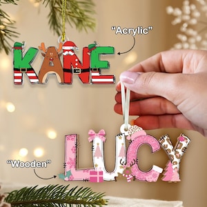 May include: Two Christmas ornaments spelling out names. One is acrylic and spells "KANE" with festive designs. The other is wooden and spells "LUCY" with pink and white patterns, including trees and bows. Both have hanging ribbons.