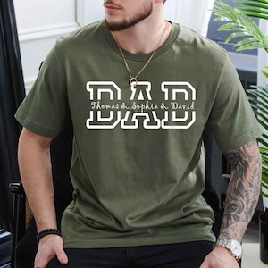 K&ouml;nnte beinhalten: Ein gr&uuml;nes T-Shirt mit einer wei&szlig;en "DAD"-Grafik. Der Text "Thomas & Sophia & David" steht unter der Grafik.
